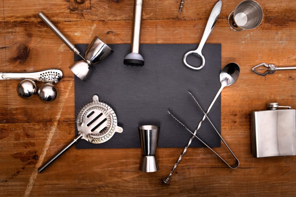 Stainless steel bar tools including a shaker strainer, jigger, muddler and bar spoon laid out on a slate board on a wooden table