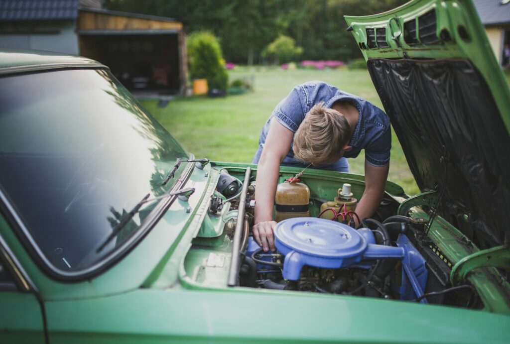 A young man leaning under the open hood of a vintage green car, working on the engine in a garden