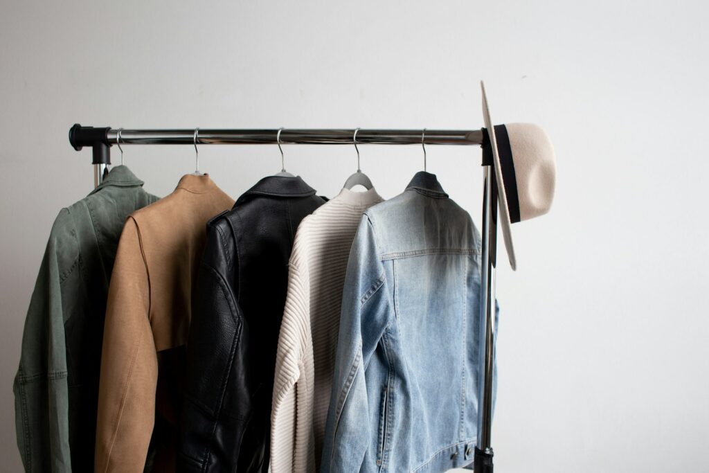A clothes rack with a green jacket, tan suede jacket, black leather jacket, cream knit and denim jacket, with a hat hanging on the end