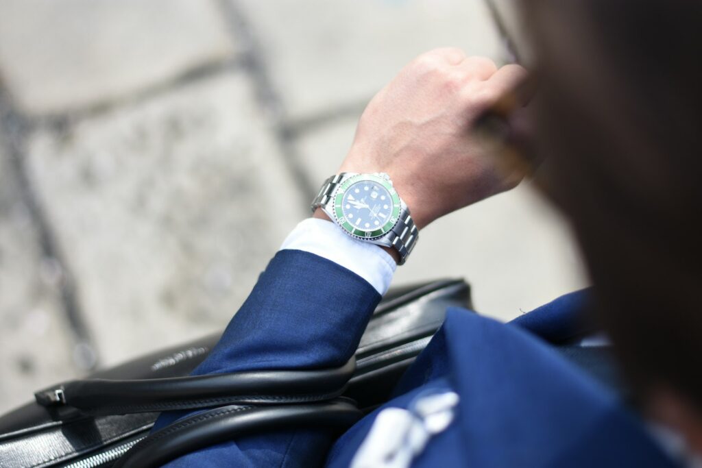 A man in a navy suit checking a steel wristwatch with a green bezel while carrying a leather bag