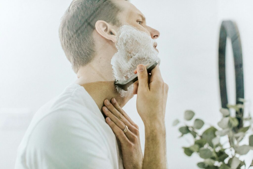A man in a white T-shirt shaving his lathered jaw in front of a mirror