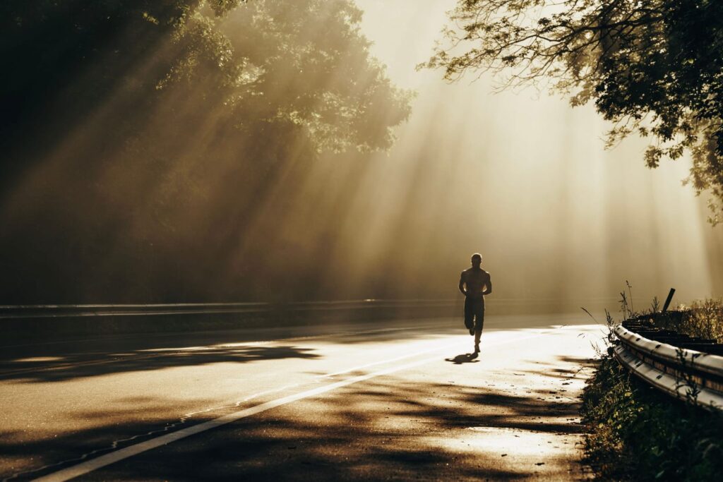 A lone runner on a quiet road through the trees with morning sunlight streaming through the branches